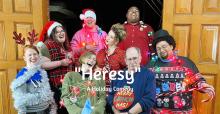 Group of people stand in doorway with text "Heresy a Holiday Comedy" centered on image
