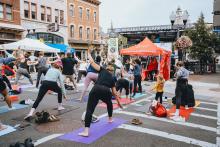 A large crowd of yoga students perform poses in front of a stage on N Pearl St in downtown Albany, New York